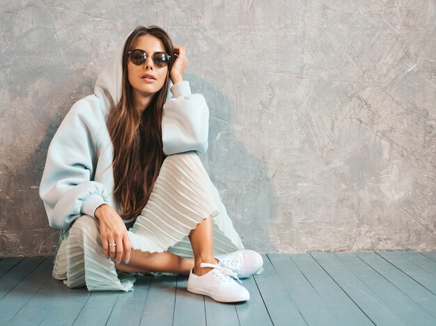 Joven hermosa mujer sonriente muestra el signo de la paz. Chica de moda en ropa casual de verano con capucha y falda. sentado en el suelo