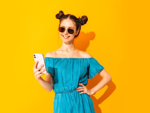 Joven hermosa mujer sonriente en overoles de jeans de verano de moda Mujer sexy despreocupada con peinado de dos cuernos posando cerca de la pared amarilla mirando la pantalla del teléfono celular Sosteniendo el teléfono inteligente y usando aplicaciones