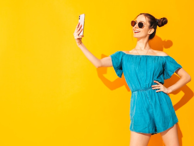 Joven hermosa mujer sonriente en overoles de jeans de verano de moda Mujer sexy despreocupada con peinado de dos cuernos posando junto a la pared amarilla en el estudio Modelo positivo divirtiéndose Aislado Tomando selfie