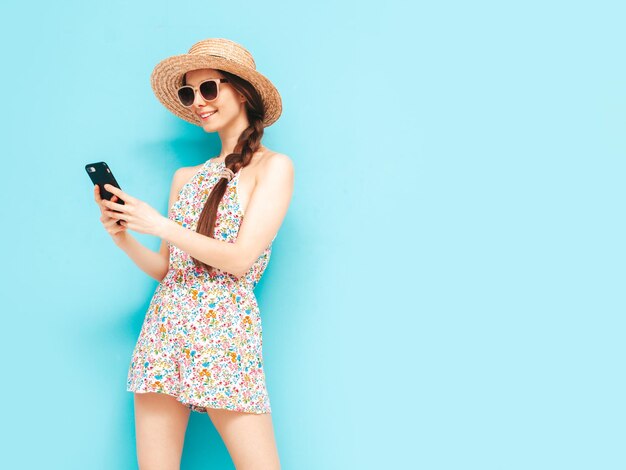 Joven hermosa mujer sonriente en overoles de verano de moda Mujer sexy con posando cerca de la pared amarilla en el estudio Modelo positivo divirtiéndose En gafas de sol y sombrero Mirando el teléfono inteligente Usando aplicaciones