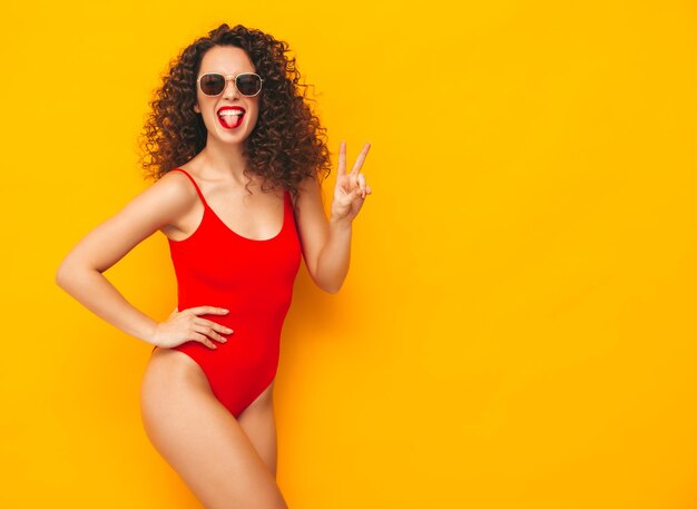 Joven hermosa mujer sonriente posando junto a la pared amarilla en el estudio Modelo sexy en traje de baño rojo Mujer positiva con peinado de rizos Feliz y alegre En gafas de sol Mostrando signo de paz