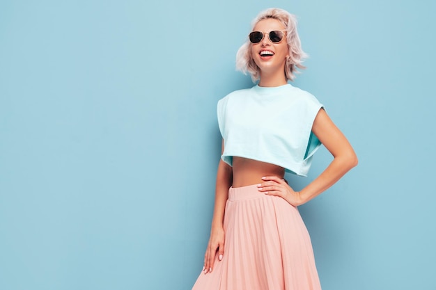 Joven hermosa mujer sonriente en ropa de verano de moda Mujer sexy despreocupada posando junto a la pared azul en el estudio Modelo rubio positivo divirtiéndose y volviéndose loco En gafas de sol