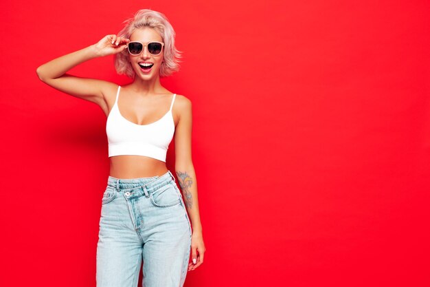 Joven hermosa mujer sonriente en ropa de verano de moda Mujer sexy despreocupada posando junto a la pared roja en el estudio Modelo rubio positivo divirtiéndose y volviéndose loco Alegre y feliz En gafas de sol