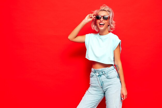 Joven hermosa mujer sonriente en ropa de verano de moda Mujer sexy despreocupada posando junto a la pared roja en el estudio Modelo rubio positivo divirtiéndose y volviéndose loco Alegre y feliz En gafas de sol