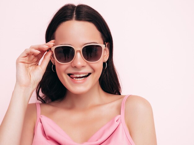 Joven hermosa mujer sonriente en ropa de verano de moda Mujer sexy despreocupada posando junto a la pared rosa en el estudio Modelo positivo divirtiéndose en el interior Quitándose las gafas de sol Muestra la lengua