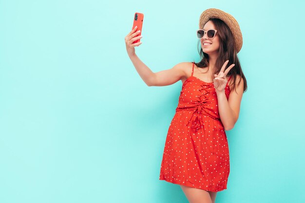 Joven hermosa mujer sonriente en vestido rojo de verano de moda Mujer despreocupada sexy posando junto a la pared azul en el estudio Modelo morena positiva divirtiéndose con sombrero y gafas de sol Tomando fotos selfie