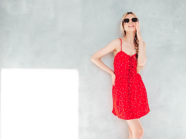 Joven hermosa mujer sonriente en vestido rojo de verano de moda Sexy mujer rubia despreocupada posando junto a la pared en el estudio Modelo positivo divirtiéndose Alegre y feliz En un día soleado