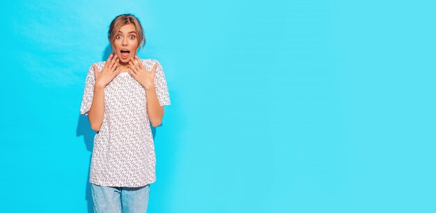 Joven hermosa mujer sorprendida y sorprendida mirando a cámara. Chica de moda en ropa casual de verano. Emoción femenina positiva expresión facial lenguaje corporal. Modelo divertido aislado sobre fondo azul