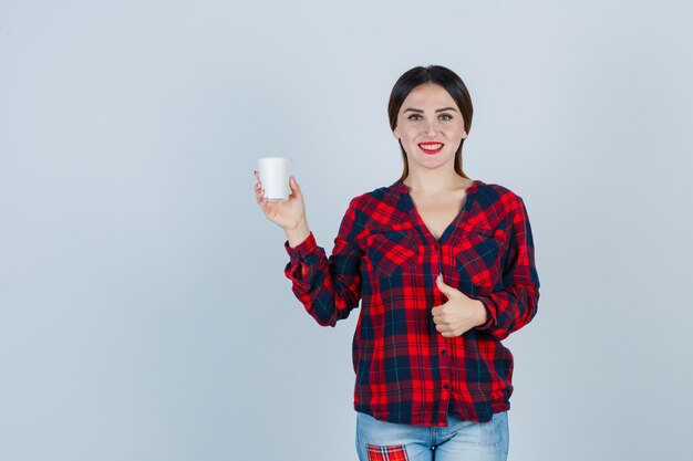 Joven hermosa mujer sosteniendo un vaso de plástico mientras muestra el pulgar hacia arriba en camisa casual, jeans y parece alegre. vista frontal.