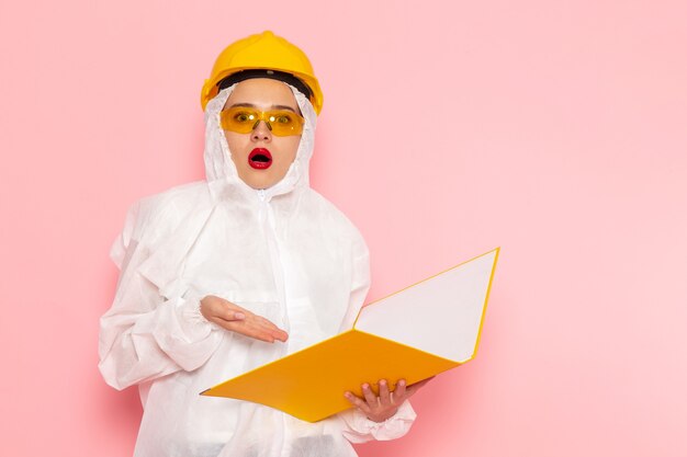 Joven hermosa mujer en traje blanco especial con casco protector sosteniendo archivo con expresión de sorpresa en rosa