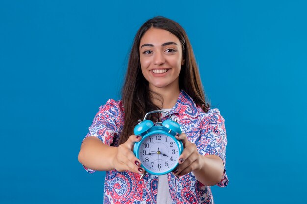 Joven hermosa mujer turista con despertador con una cara feliz y sonriendo sobre pared azul aislado