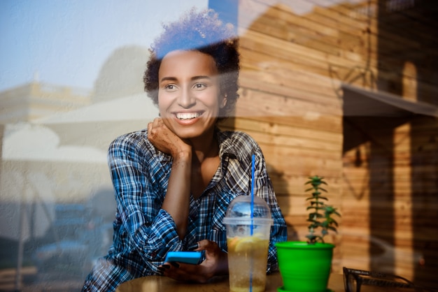 Joven hermosa niña africana sonriendo, sosteniendo el teléfono, sentado en la cafetería. Disparo desde afuera.