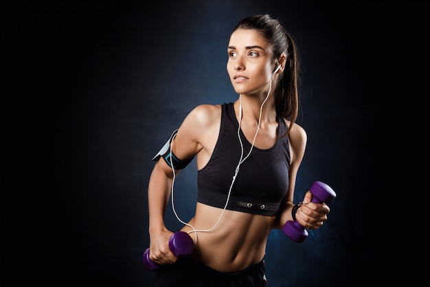 Joven hermosa niña deportiva entrenamiento con pesas sobre la pared oscura.