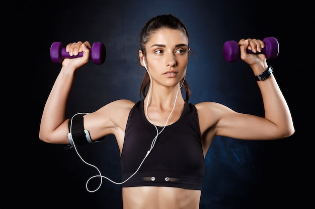 Joven hermosa niña deportiva entrenamiento con pesas sobre la pared oscura.