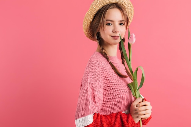 Joven hermosa niña sonriente con dos trenzas en suéter y sombrero de paja sosteniendo un tulipán en la mano mirando soñadoramente a la cámara mientras pasa tiempo sobre fondo rosa