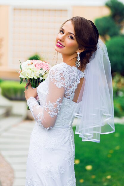 Joven hermosa novia feliz en vestido de novia blanco elegante con ramo posando al aire libre en el parque.