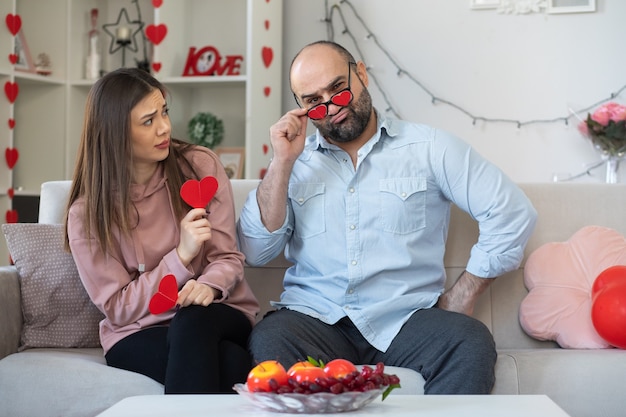 Joven hermosa pareja disgustó a hombre con gafas divertidas y mujer confundida con corazones hechos de cartón celebrando el día internacional de la mujer sentada en un sofá en la sala de estar luminosa