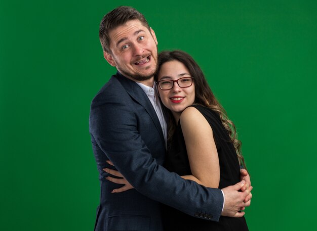 Joven hermosa pareja feliz y alegre hombre y mujer abrazando felices enamorados juntos celebrando el día de San Valentín de pie sobre la pared verde