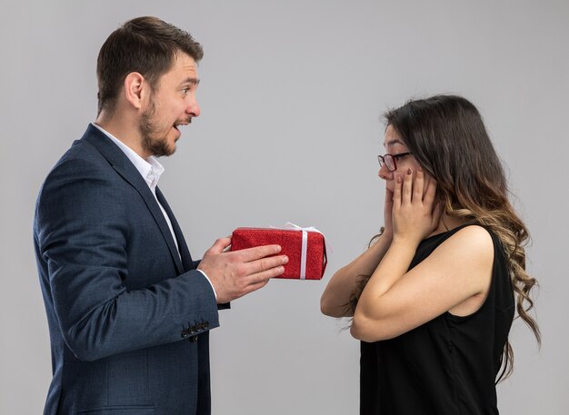 Joven hermosa pareja feliz dando un regalo para su encantadora novia feliz enamorado celebrando el día de San Valentín de pie sobre una pared blanca