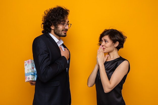 Joven hermosa pareja feliz escondido presente de su novia sorprendida y confundida celebrando el día internacional de la mujer el 8 de marzo de pie sobre fondo naranja