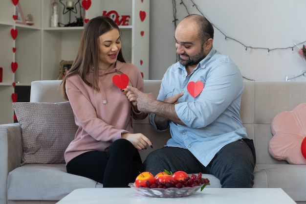 Joven hermosa pareja feliz hombre y mujer con corazones hechos de cartón sonriendo divirtiéndose juntos celebrando el día de San Valentín sentados en un sofá en la sala de luz