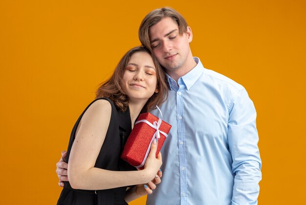 Joven hermosa pareja feliz hombre y mujer con presente en manos sonriendo alegremente abrazando felices enamorados juntos celebrando el día de San Valentín de pie sobre la pared naranja