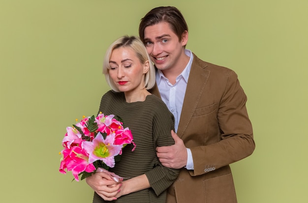 Joven hermosa pareja feliz hombre y mujer con ramo de flores abrazándose felices enamorados juntos sonriendo celebrando el día internacional de la mujer de pie sobre la pared verde