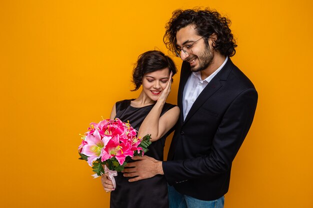 Joven hermosa pareja feliz hombre y mujer con ramo de flores sonriendo alegremente abrazando feliz enamorado celebrando el día internacional de la mujer el 8 de marzo de pie sobre fondo naranja