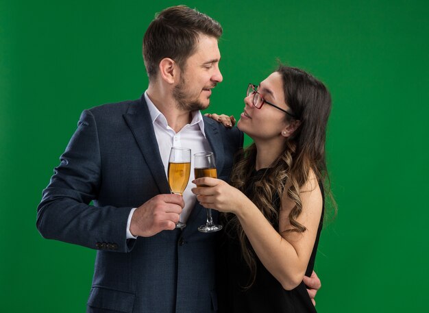 Joven hermosa pareja feliz hombre y mujer sonriente con copas de champán abrazándose mirando el uno al otro feliz en el amor juntos celebrando el día de San Valentín de pie sobre la pared verde