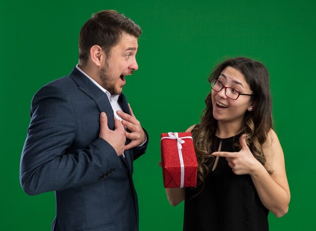 Joven hermosa pareja feliz recibiendo un regalo de su novia feliz enamorado juntos celebrando el día de San Valentín de pie sobre la pared verde