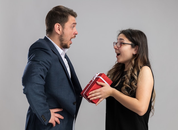 Joven hermosa pareja feliz y sorprendido mirando a su encantadora novia con un regalo para él celebrando el día de San Valentín de pie sobre una pared blanca