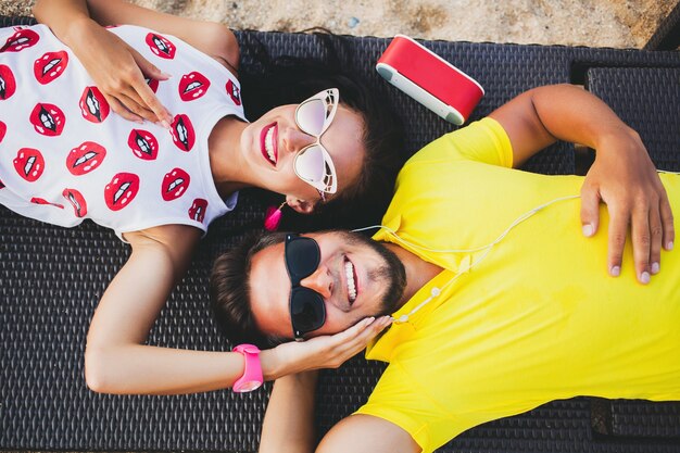 Joven hermosa pareja hipster enamorada, acostado abrazando, escuchando música, gafas de sol, traje elegante, vacaciones de verano, divirtiéndose, sonriendo, feliz, colorido, vista desde arriba