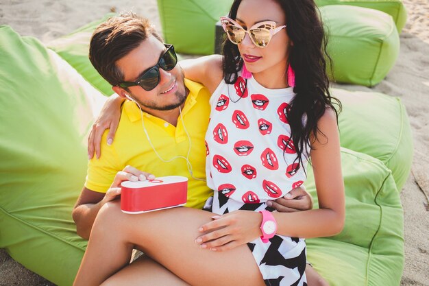 Joven hermosa pareja hipster enamorada sentada en la playa, escuchando música, gafas de sol, atuendo elegante, vacaciones de verano, emoción colorida y positiva