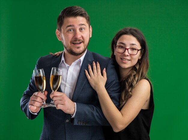 Joven hermosa pareja hombre feliz con copas de champán y mujer sonriente abrazando feliz enamorado juntos celebrando el día de San Valentín de pie sobre la pared verde