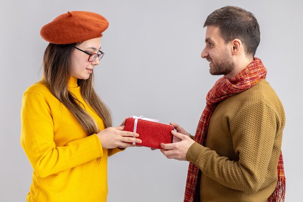 Joven hermosa pareja hombre feliz dando un regalo a su novia sonriente en boina feliz enamorado juntos celebrando el día de San Valentín de pie sobre una pared blanca