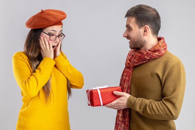 Joven hermosa pareja hombre feliz dando un regalo a su novia sonriente y sorprendida en boina feliz enamorado juntos celebrando el día de San Valentín de pie sobre fondo blanco