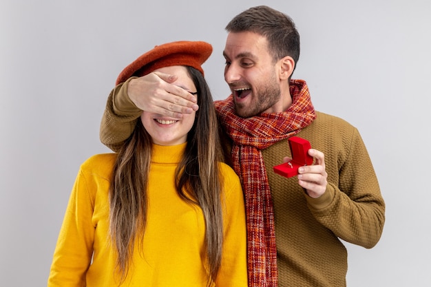Joven hermosa pareja hombre feliz haciendo propuesta con anillo de compromiso en caja roja a su novia asombrada en boina cubriendo sus ojos durante el día de San Valentín de pie sobre una pared blanca