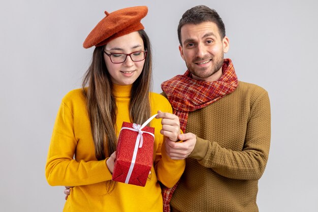 Joven hermosa pareja hombre feliz y mujer sonriente en boina sosteniendo presente feliz enamorado juntos celebrando el día de San Valentín de pie sobre una pared blanca