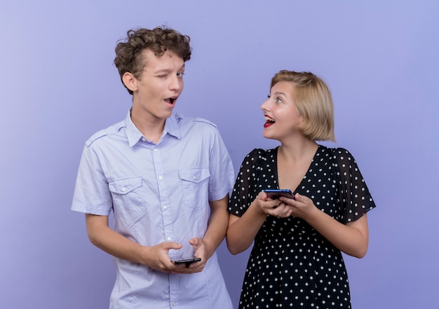 Joven hermosa pareja hombre y mujer feliz y positivo sosteniendo teléfonos inteligentes sonriendo y guiñando un ojo de pie sobre la pared azul