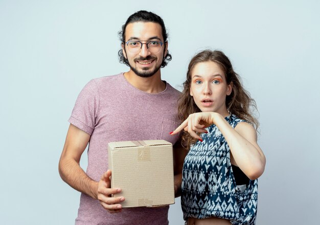 Joven hermosa pareja hombre y mujeres, hombre sosteniendo el paquete de caja sonriendo mientras su novia apunta con el dedo a la caja sorprendida sobre fondo blanco.