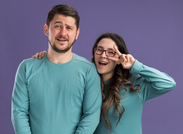 Joven hermosa pareja en ropa casual azul hombre y mujer feliz y alegre sonriendo divirtiéndose mostrando v-sign feliz en el amor de pie sobre la pared púrpura