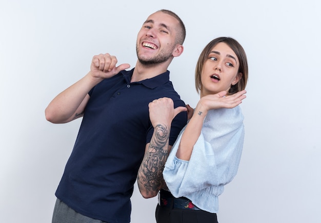 Joven hermosa pareja en ropa casual hombre y mujer hombre feliz y alegre apuntando con los pulgares a su novia sonriendo alegremente de pie sobre la pared blanca