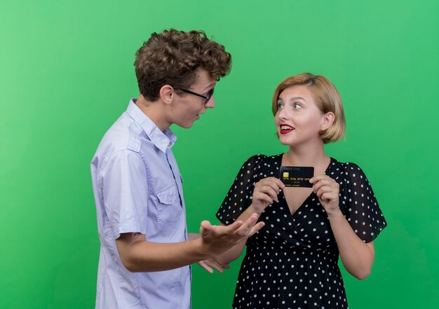 Joven hermosa pareja sorprendió a hombre mirando a la novia feliz que sostiene la tarjeta de crédito de pie sobre la pared verde