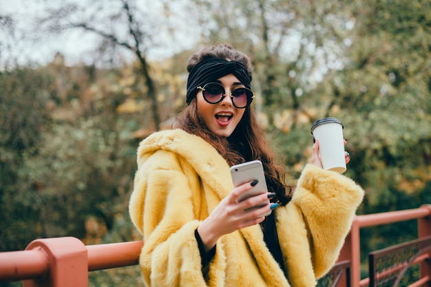 La joven hermosa usa un teléfono inteligente en la calle, navega en internet y toma café.