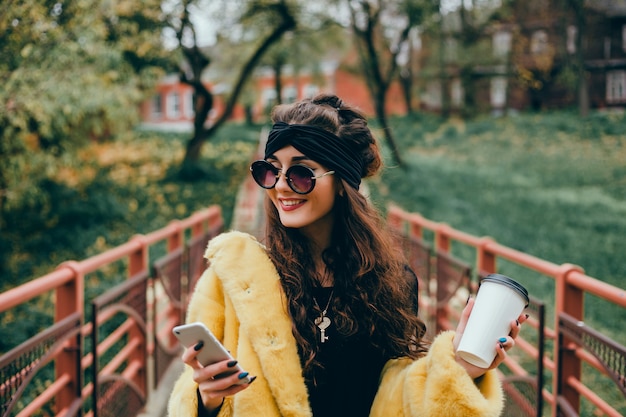 La joven hermosa usa un teléfono inteligente en la calle, navega en internet y toma café.