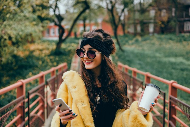 La joven hermosa usa un teléfono inteligente en la calle, navega en internet y toma café.