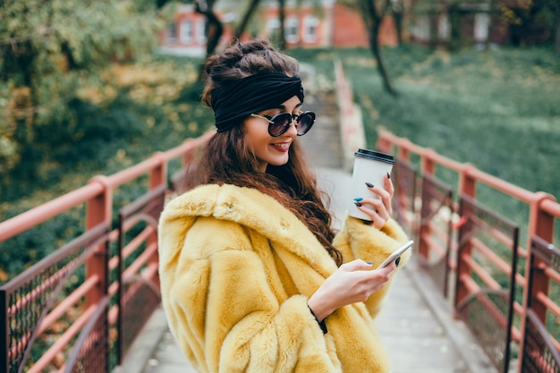 La joven hermosa usa un teléfono inteligente en la calle, navega en internet y toma café.