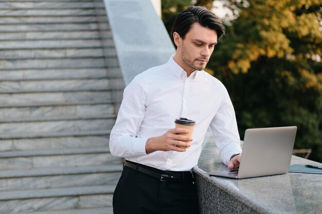 Joven hermoso y serio hombre moreno barbudo con camisa blanca sosteniendo una taza de café para ir de la mano trabajando cuidadosamente en una laptop en el parque de la ciudad solo