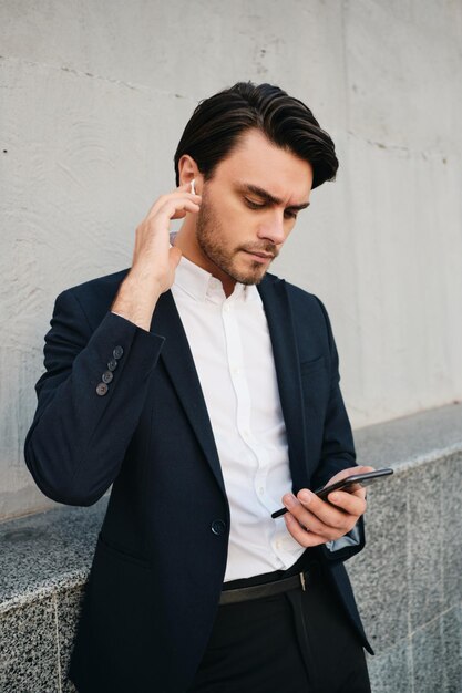 Joven hermoso y serio hombre moreno barbudo con camisa blanca y traje clásico con auriculares inalámbricos cuidadosamente usando el teléfono celular mientras está parado cerca de una pared gris en la calle