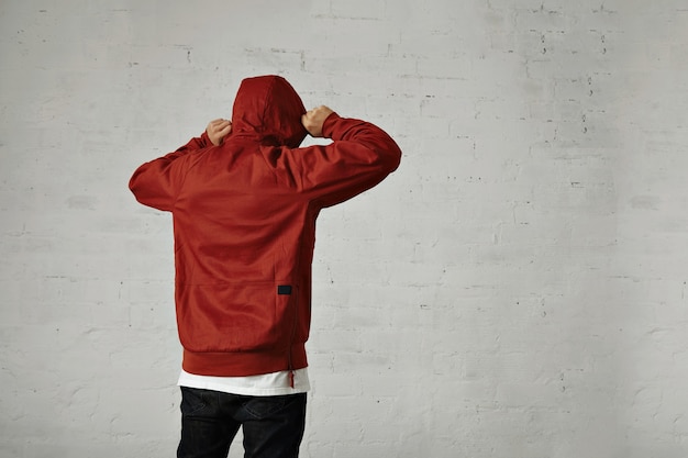 Un joven hipster ajusta la capucha de su parka rojo pardusco, vista posterior, retrato en estudio con paredes blancas